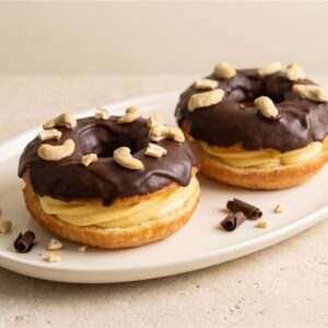 A full-size donut layered with custard and finished with chocolate and cashew pieces. Serving presentation.